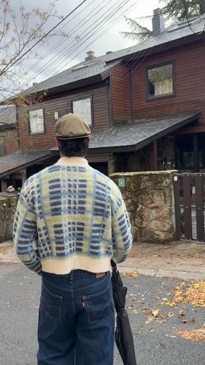Pull en tricot bleu clair et vert pâle avec un motif à carreaux, porté sur un jean en denim foncé. Une personne tient un parapluie noir.