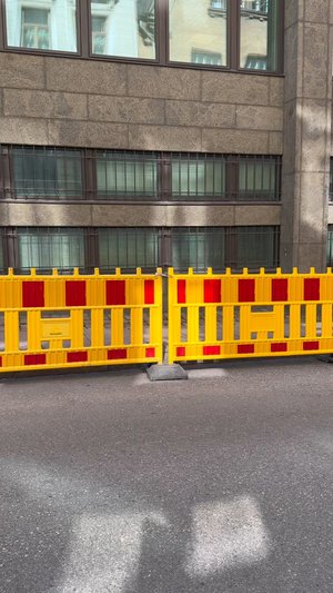 Barrière en plastique jaune avec des bandes réfléchissantes rouges, montée sur une base en béton gris, contre un mur de pierre et des fenêtres d'un bâtiment.