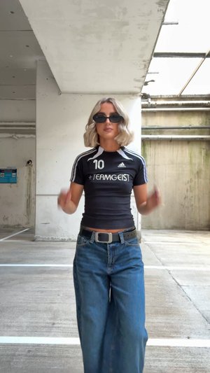 Haut noir à manches courtes avec des rayures blanches et le logo "TEAMGEIST", associé à un jean en denim bleu à jambes larges et une ceinture noire, portant des lunettes de soleil sombres.