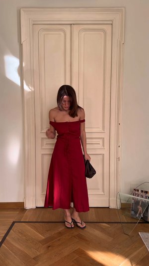 Rotes schulterfreies Kleid mit Knopfdetails und Bindeband in der Taille, das eine fließende Silhouette bietet. Schwarze Handtasche und Flip-Flops vervollständigen den Look.