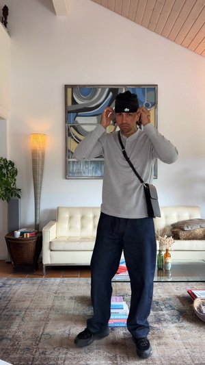 Szürke hosszúujjú póló, sötétkék bő nadrág, fekete sapka és keresztpántos táska, egy mintás szőnyegen áll egy világos szobában.