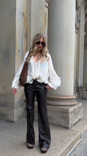 White ruffled blouse with long sleeves, high-waisted black leather flared trousers, and a brown shoulder bag. Brown wedge shoes complete the outfit.