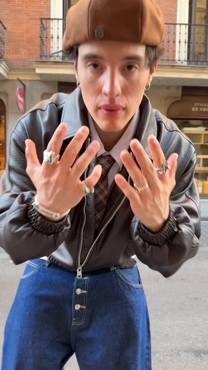 Béret marron, bagues argentées aux mains, veste en cuir noire avec une fermeture éclair, et pantalon en denim bleu avec détail de bouton. Chemise fine à rayures en dessous.