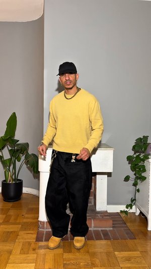 Pull jaune, pantalon noir ample, bottes couleur tan ; porte une casquette noire et un collier chaîne ; cadre intérieur avec des plantes en pot et des murs neutres.