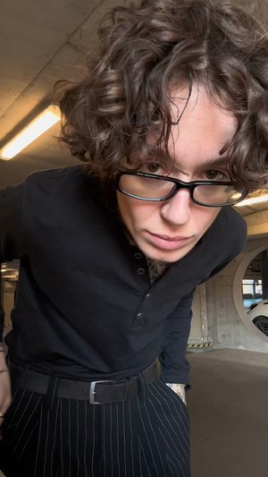 Melna garām piedurknēm krekls, svītrainas bikses un melns josta. Viļņaini pusgarie mati un brilles, betonā stāvvietā.