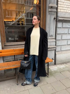 Cappotto lungo nero, maglione crema, jeans blu larghi con risvolti, handbag nera con dettagli dorati e sneakers multicolori. Ambiente urbano.