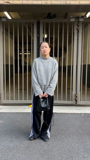 Pull gris en tricot avec manches larges, pantalon de survêtement noir avec des bandes blanches, et un petit sac noir. Debout devant une zone clôturée.