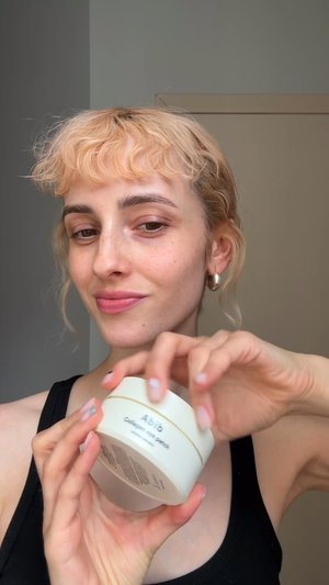 Ronde, witte huidverzorgingscontainer met gouden accent; gelabeld 'Abib Collagen Gel Patch.' Vastgehouden in een hand, met een textuur achtergrond.