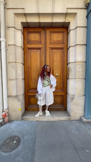 White oversized shirt, green patterned top, white shorts, white sneakers. Silver handbag, wooden door backdrop, gray pavement.
