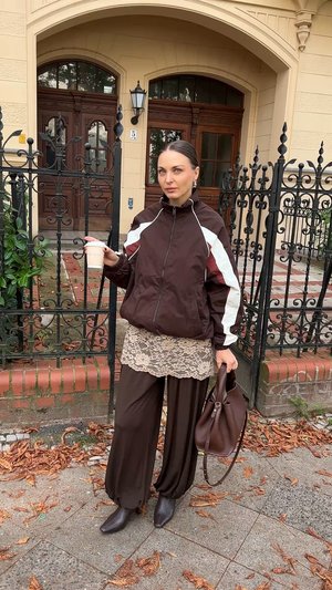 Barna cipzáras dzseki fehér és bordó díszítéssel, csipkés felső, széles barna nadrág, egy bögre a kezében és egy barna táska.