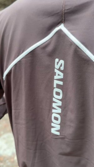 Brown athletic shirt with a white zipper and the word "SALOMON" printed vertically in white along the side. Outdoor setting in soft focus.