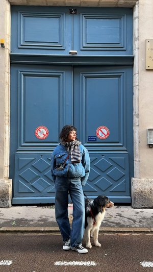 A person in a blue denim outfit with a striped scarf stands beside a dog in front of blue double doors, with no-parking signs visible.