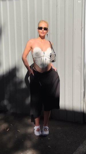 Rövid szőke hajú nő napszemüvegben, fehér, formázott melltartóban, fekete, bő szárú nadrágban, fehér cipővel és egy kis táskával áll a szabadban.