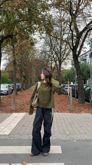 Pull oversize vert olive, pantalon ample noir avec des bandes blanches, et un sac à main à motif. Feuilles d'automne et arbres en arrière-plan.