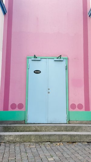 Világoskék dupla ajtók zöld keretben, rózsaszín, függőleges csíkokkal és körökkel díszített fal előtt. Kőlépcsők vezetnek az ajtóhoz.