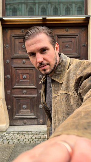 Une veste en cuir marron avec une texture montrant des signes d'usure, portée sur un pull en maille gris, sur fond d'une porte en bois avec des accents métalliques ronds.