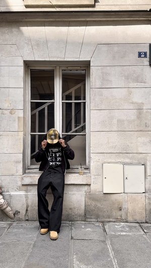 Pantalons larges noirs, un t-shirt graphique noir et une casquette à carreaux jaunes avec un logo. Debout devant un mur en pierre et une fenêtre.