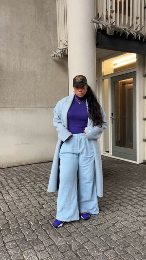 Világoskék túlméretezett kabát, lila felső és világoskék bőszárú nadrág. Fekete sapka és kék cipők részletekkel. Texturált macskaköves háttér.