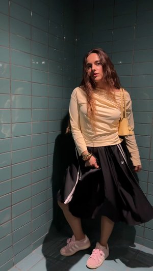 Haut à manches longues jaune avec détail drapé, jupe noire à rayures blanches, et baskets roses. L'arrière-plan présente des carreaux turquoise.