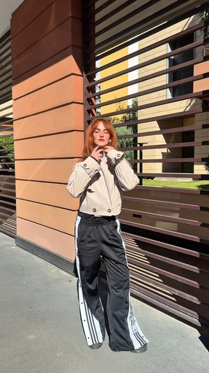 Beige cropped Jacke mit großen Knöpfen, kombiniert mit schwarzen Hosen mit weißen Seitenstreifen. Vor einer strukturierten Wand und einem lamellenartigen Hintergrund.