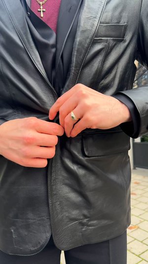 Black leather blazer with a slim fit, featuring a notch lapel and a single pocket. Hands adjusting the button, displaying a ring with a green stone.
