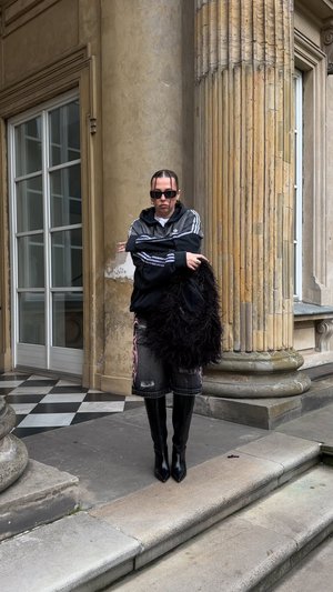 Bottes noires en cuir montant jusqu'aux genoux, veste zippée gris et noir, short en denim déchiré avec des motifs roses, tenant un châle noir duveteux.