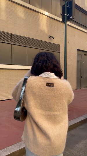 Beige fuzzy trui met een lage halslijn, voorzien van een donker patchlabel, gecombineerd met een glanzende zwarte schoudertas, gedragen met lichtblauwe jeans.