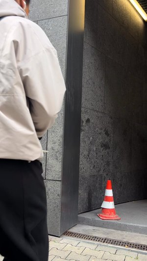 Personne en veste beige se tenant près d'un mur en pierre grise avec un cône de circulation orange et blanc sur un trottoir surélevé dans une ruelle sombre.