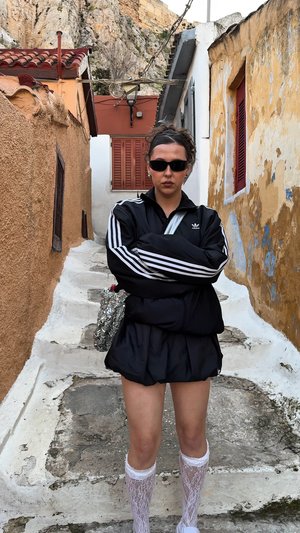 Zwarte sportjack met witte strepen, bijpassende zwarte rok, witte kanten kniekousen en een zonnebril, tegen een stenen steeg.