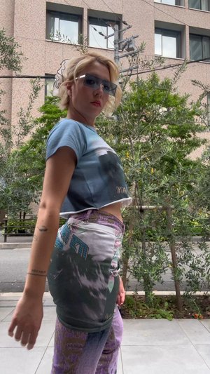 Kék, logóval díszített, fixált grafikus póló, mintás lila és fehér nadrággal párosítva. A viselője egy zöld cserje háttér előtt pózol.