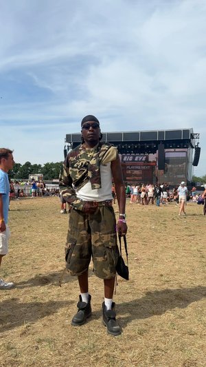 Chemise et short à motif camouflage, bottes de combat, lunettes de soleil et un bandana noir ; debout sur l'herbe avec une scène en arrière-plan.