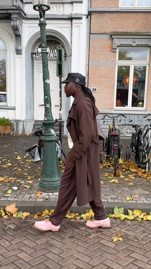 Manteau en cuir marron avec larges revers, pantalon assorti et chaussures chunky rose pâle ; marchant sur un trottoir en pavés jonché de feuilles.