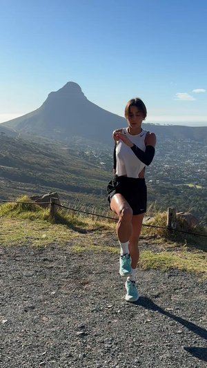 Atletka ve bezrukávovém bílém tričku a černých šortkách provádí běžecké cvičení na štěrku, s horským pozadím pod jasnou modrou oblohou.