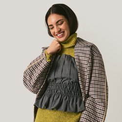 Mladá žena se usmívá, má na sobě kostkovaný blejzr přes zelený rolák a šedý top s volánky, pruhované šedé kalhoty, stojí před jednobarevným pozadím.