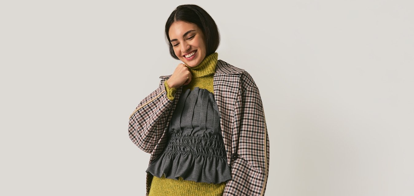 Mladá žena se usmívá, má na sobě kostkovaný blejzr přes zelený rolák a šedý top s volánky, pruhované šedé kalhoty, stojí před jednobarevným pozadím.