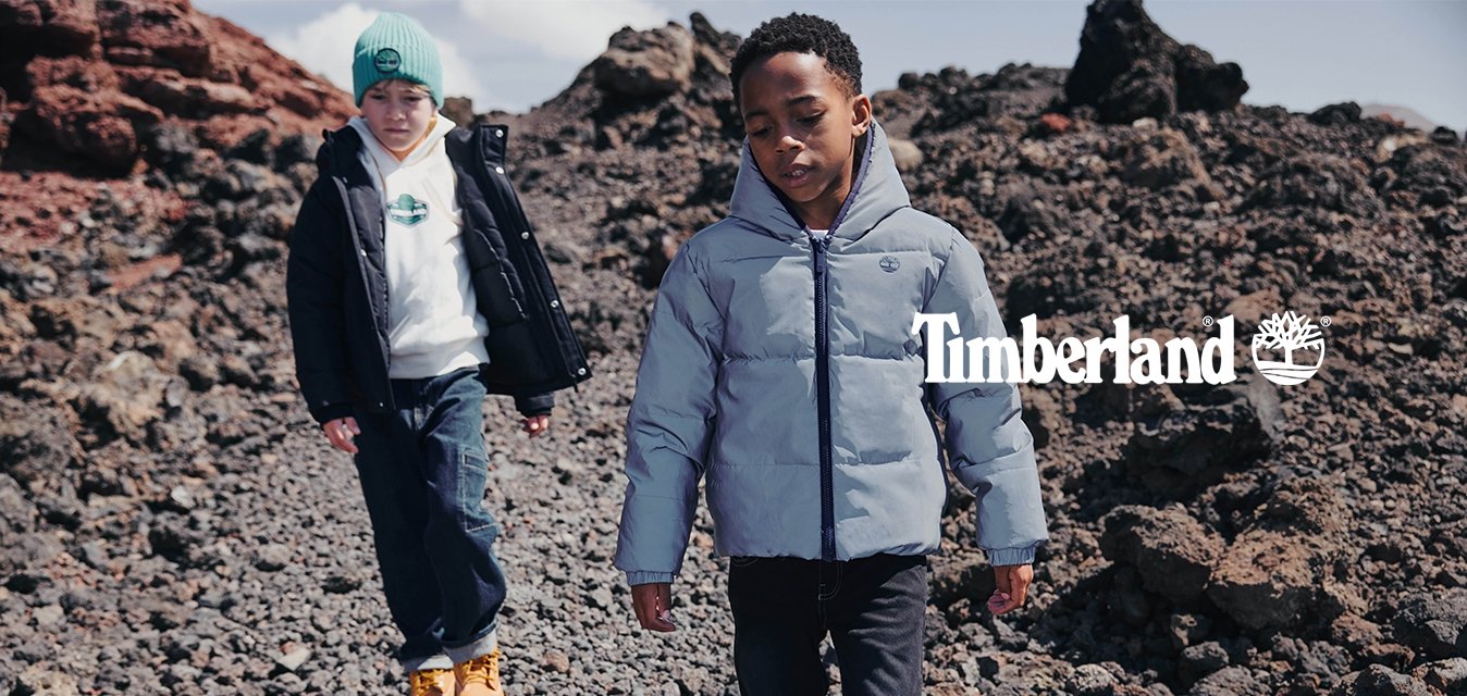 Dva chlapci v zimních bundách kráčejí po kamenitém terénu s vulkanickými kameny pod částečně oblačnou oblohou.