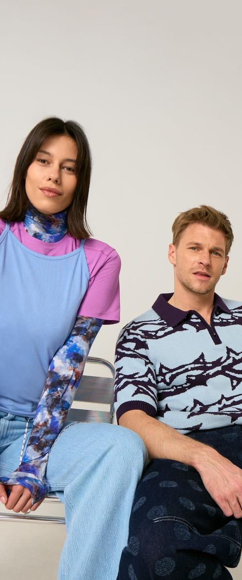 Jeune femme et homme assis, tous deux portant des hauts à motifs et des pantalons décontractés, sur un fond clair uni.