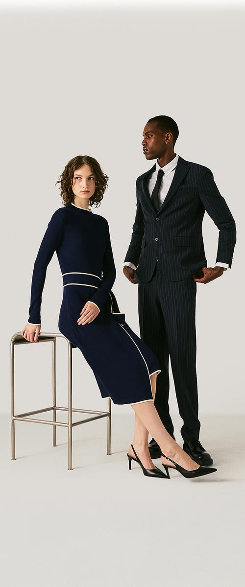 Woman in navy dress with white trim sits on a metal stool, man in dark pinstripe suit stands beside her against plain background.