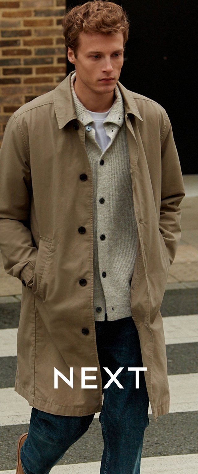 Khaki płaszcz trenczowy z zapięciem na guziki, zestawiony z jasnoszarym kardiganem. Ciemnoniebieskie dżinsy dopełniają stylizację, a tło stanowi subtelne przejście dla pieszych.