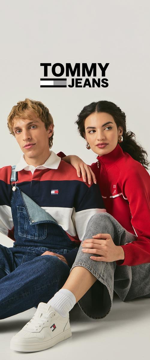 Modelo con un top rojo con cremallera y pantalones vaqueros de peto sentado junto a otro con un polo a rayas. Ambos llevan zapatillas blancas. Logo "TOMMY JEANS" arriba.