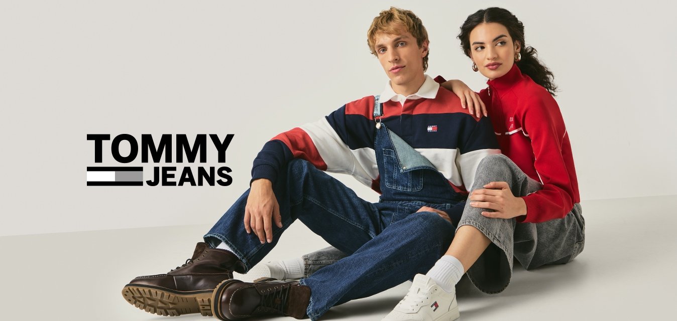 Muž v pruhovaném tričku a džínových laclích sedí vedle ženy v červeném tričku s dlouhým rukávem a šedých kalhotách na jednobarevné bílé podlaze.