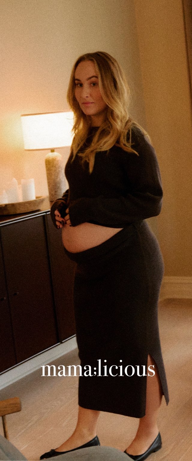 Zwangere jurk in donkere stof met lange mouwen, aansluitend ontwerp en een zijsplit. Gekenmerkt door een vrouw die binnen poseert met een zachte lamp in de buurt.