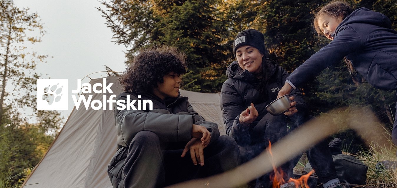 Tři lidé sedí kolem táborového ohně s béžovým stanem v pozadí. Mají na sobě zateplené bundy a boty, obklopeni trávou a stromy.