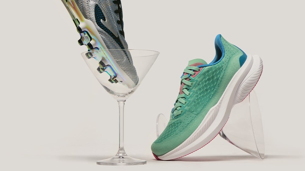Grüner Sportschuh mit texturiertem Mesh-Obermaterial, blauen Akzenten, pinkfarbenen Schnürsenkeln und weißer Sohle, neben einem transparenten Martiniglas.
