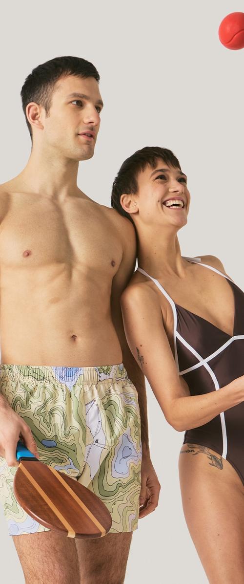 Jeune homme et femme en maillots de bain jouant au paddle ball, l'homme tenant une raquette en bois, balle rouge en suspension sur un fond uni.