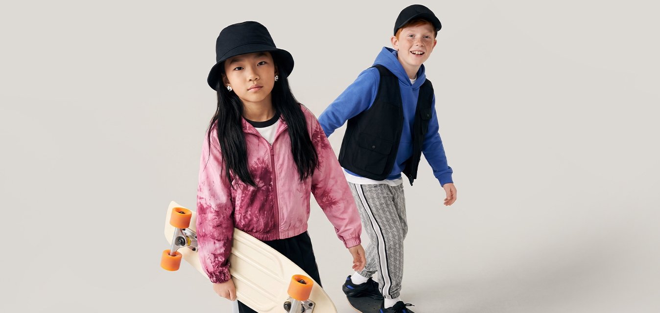 Dvě děti na skateboardu; dívka drží skateboard, chlapec jezdí na desce, oba mají na sobě běžné sportovní oblečení na jednoduchém světlém pozadí.