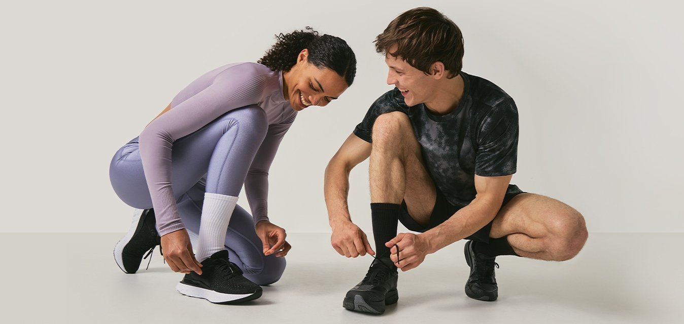 Dva lidé v sportovním oblečení se skrčí a zavazují si běžecké boty na jednoduchém světle zbarveném pozadí.
