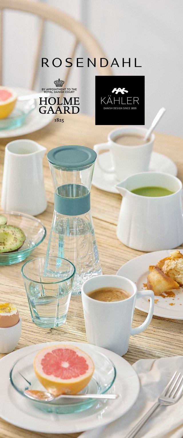 Glass pitcher with teal accent, white porcelain mugs, clear plates, and various food items including grapefruit and croissants on a wooden table.