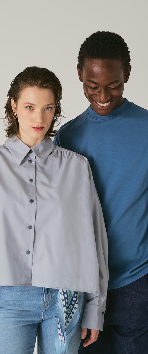 Helles graues, kurz geschnittenes Shirt mit Knopfverschluss und puffigen Ärmeln, kombiniert mit einem blauen Langarmoberteil. Blaue Bänderdetails sichtbar.
