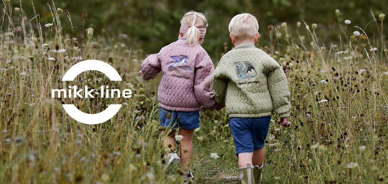 Dva děti, držící se za ruce, jdou po úzké stezce mezi vysokou trávou a divokými květinami směrem k zeleným kopcům pod zataženou oblohou.